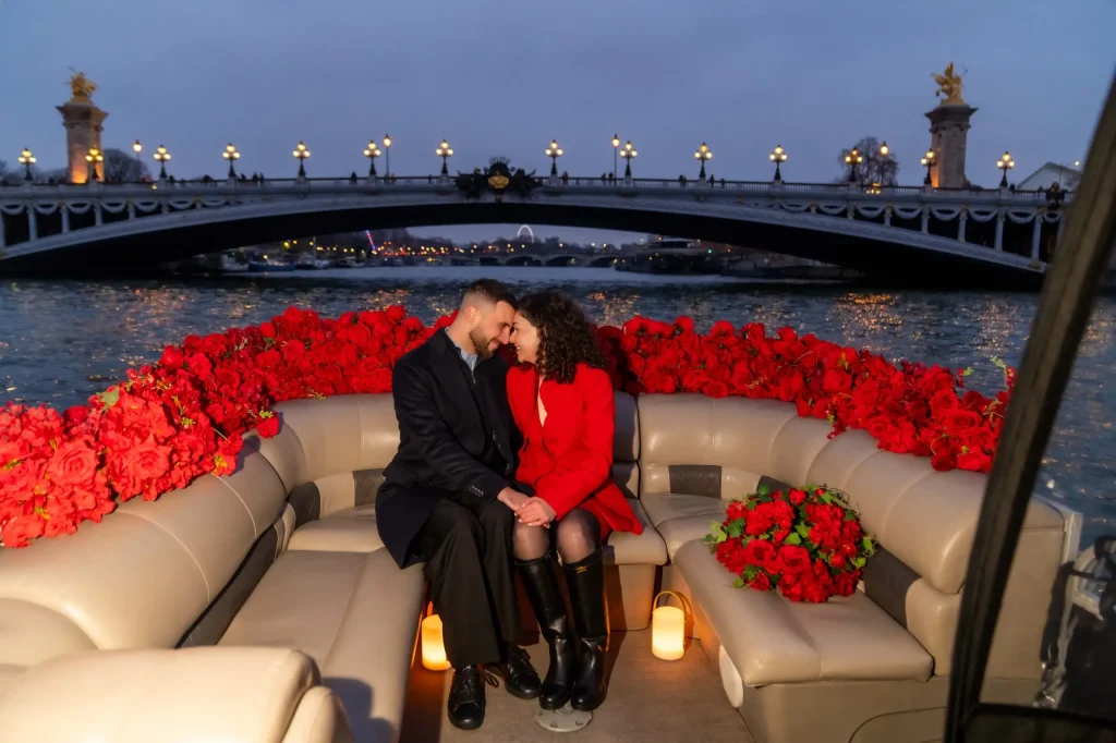 Couple sharing an intimate moment on a private Seine boat proposal in Paris, surrounded by red roses and candles with the illuminated Pont Alexandre III bridge at dusk